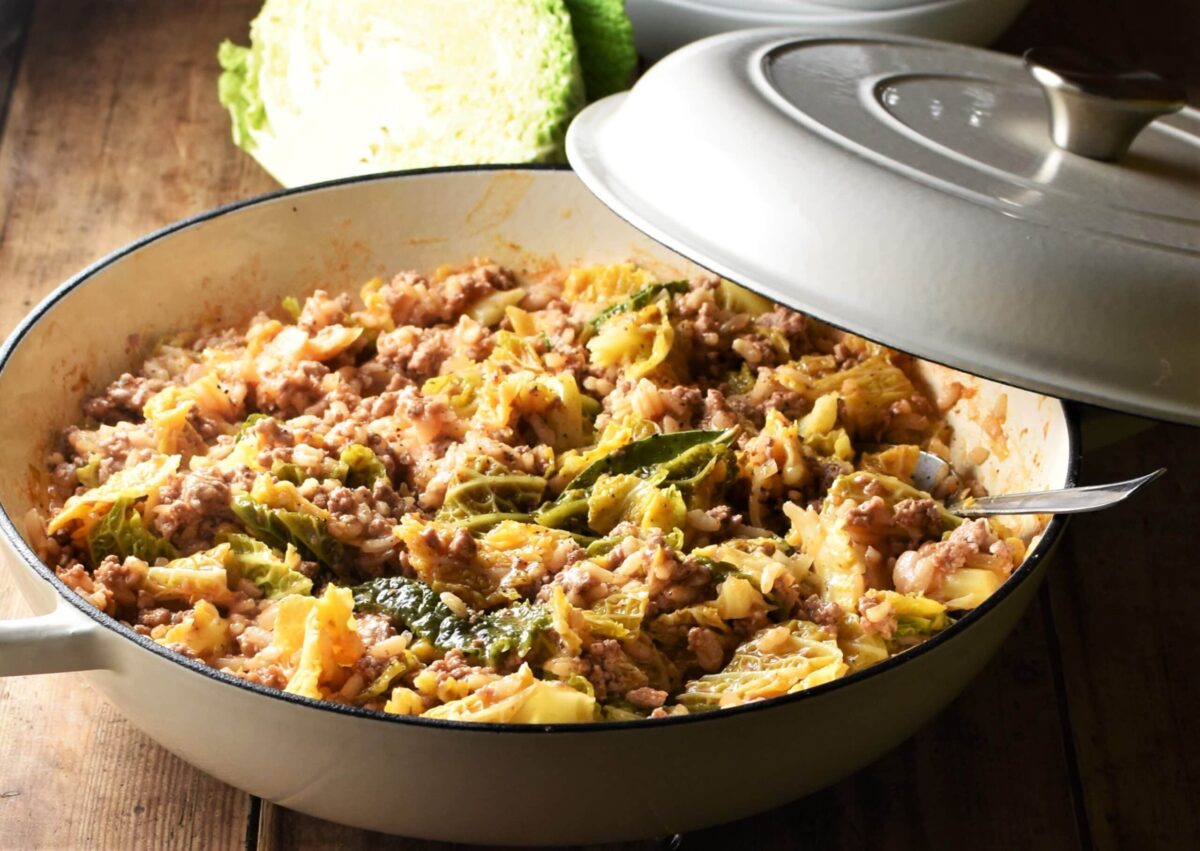 Side view of cabbage, meat and rice mixture in large shallow white dish with spoon and partly closed lid and cut cabbage in background.