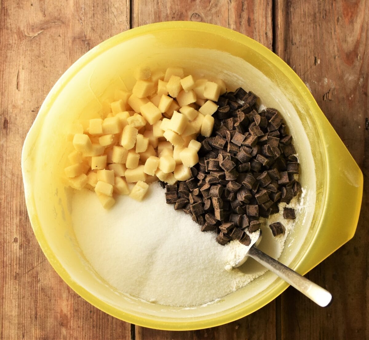Marzipan chunks, chocolate chunks and dry ingredients in yellow bowl with spoon.
