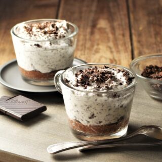 Side view of 2 cheesecake cups with chocolate shavings and spoons in background.