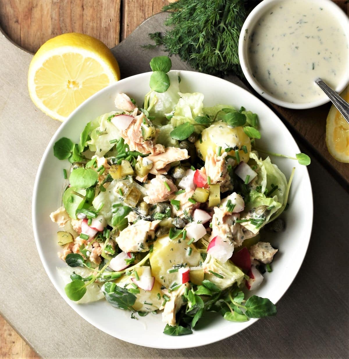 Top down view of salmon, potato and vegetable salad in white bowl with lemon and dressing in background.