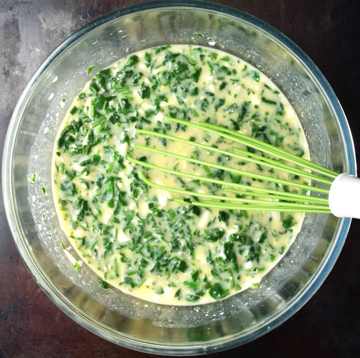 Egg mixture with spinach and whisk  in glass bowl.