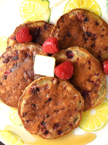 Top down view of 5 raspberry pancakes with fresh raspberries on top of yellow plate.