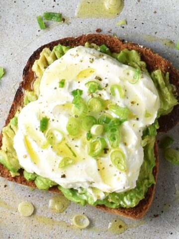 Top down view of avocado feta toast with scallions and honey on speckled plate.